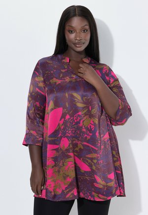 Woman wearing a bright floral tunic with 3/4 sleeves, standing against a plain white background, touching the neckline with one hand.