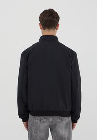 Giacca bomber nera con colletto alto, polsini e orlo a coste; texture liscia, colore uniforme, senza elementi hardware visibili o fantasie.