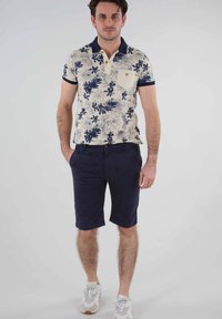 Polo à motifs floraux en crème avec des accents bleu marine et une poche, associé à un short bleu marine foncé et des baskets blanches avec des accents gris.