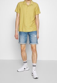 Camisa de manga corta con cuello en amarillo claro, combinada con pantalones cortos de denim remangados. Zapatillas blancas con acentos negros y calcetines a rayas.