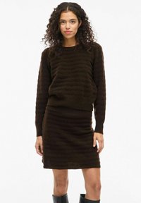 Una donna con capelli ricci indossa un maglione a maglia testurizzata marrone scuro e una gonna abbinata, abbinati a stivali neri fino al ginocchio, su uno sfondo neutro.