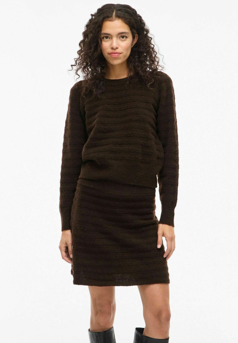 Una donna con capelli ricci indossa un maglione a maglia testurizzata marrone scuro e una gonna abbinata, abbinati a stivali neri fino al ginocchio, su uno sfondo neutro.
