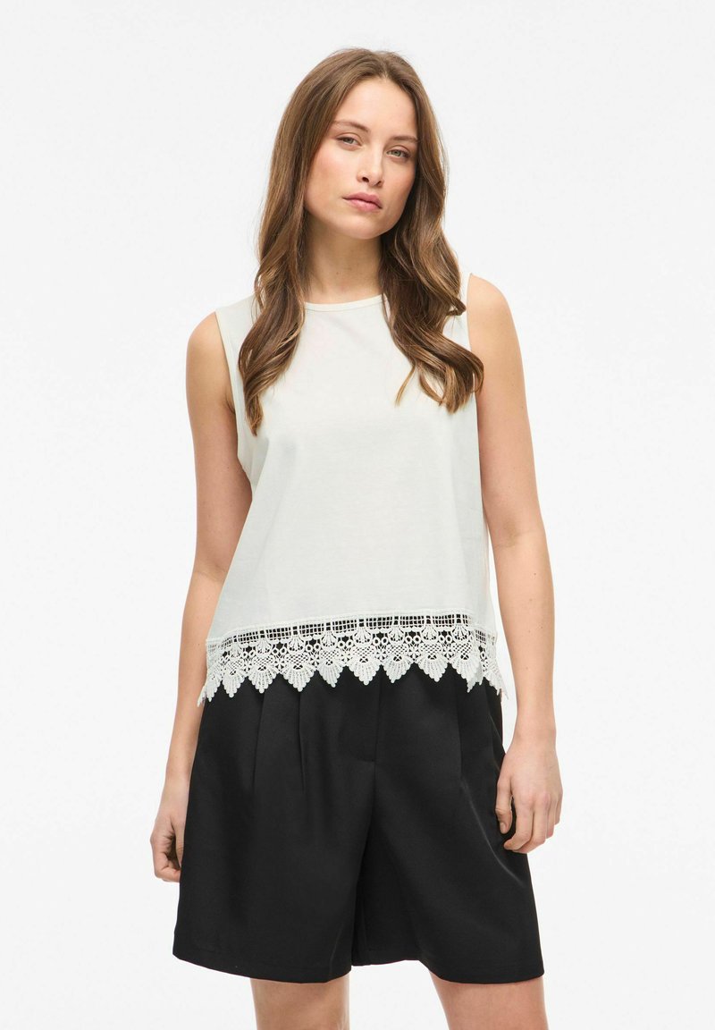 Jeune femme aux longs cheveux bruns portant un haut blanc sans manches avec un ourlet en dentelle et un short noir ample, debout devant un fond blanc.
