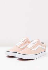 Ljusrosa mockasneakers med vita snören, en vit sidostripe och en svart gummisul. Har en låg toppdesign och en slät yta.