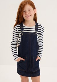 Abito in denim blu scuro con due tasche anteriori; indossato su una camicia a maniche lunghe a righe bianche e nere.