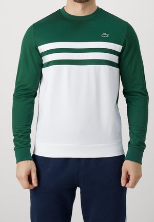 Mężczyzna noszący bluzę z długim rękawem z zieloną górną częścią, białą dolną częścią, dwoma białymi paskami na klatce piersiowej i małym zielonym logo krokodyla.