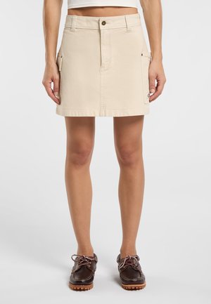 Beige cotton mini skirt with front pockets, a button closure, and a straight cut. Worn with brown lace-up shoes. Smooth texture.
