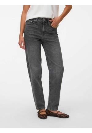 Personne portant un jean noir délavé taille haute, un haut blanc et des chaussures plates tissées marron, debout avec une main dans la poche.