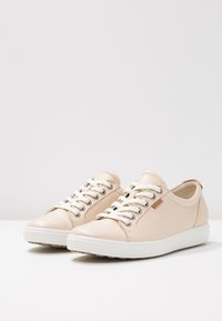 Light beige leather sneakers with white laces, silver eyelets, and a low-profile rubber sole. Features a tan logo accent.