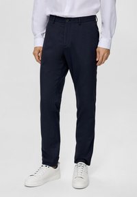Pantaloni su misura blu navy in tessuto liscio e testurizzato, con chiusura a bottone e tasche, abbinati a sneakers bianche.