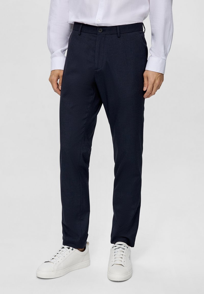 Pantaloni su misura blu navy in tessuto liscio e testurizzato, con chiusura a bottone e tasche, abbinati a sneakers bianche.