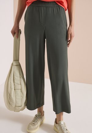 Personne portant un pantalon large gris foncé et des baskets beiges, tenant un sac beige à structure souple d'une main.