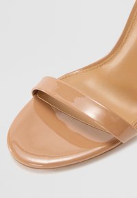 Nude patent leather aukštakulniai sandalai su blizgiu dirželiu, suapvalintu atviru pirštu, lygiu paviršiumi ir subtiliomis siuvimo detalėmis.