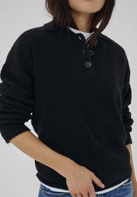 Frau trägt einen schwarzen, flauschigen, geknöpften Pullover über einem weißen Hemd, die Ärmel hochgekrempelt, und blaue Jeans vor einem neutralen Hintergrund.