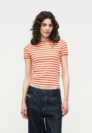 Femeie cu păr creț și închis la culoare, purtând un tricou cu mâneci scurte în dungi portocalii și albe și blugi bleumarin, stând în picioare pe un fundal simplu.