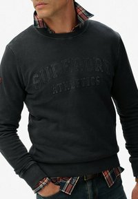 Hombre que lleva un sudadera oscura con el texto "Superdry Athletics", una camisa de cuadros debajo y jeans oscuros con las manos en los bolsillos.