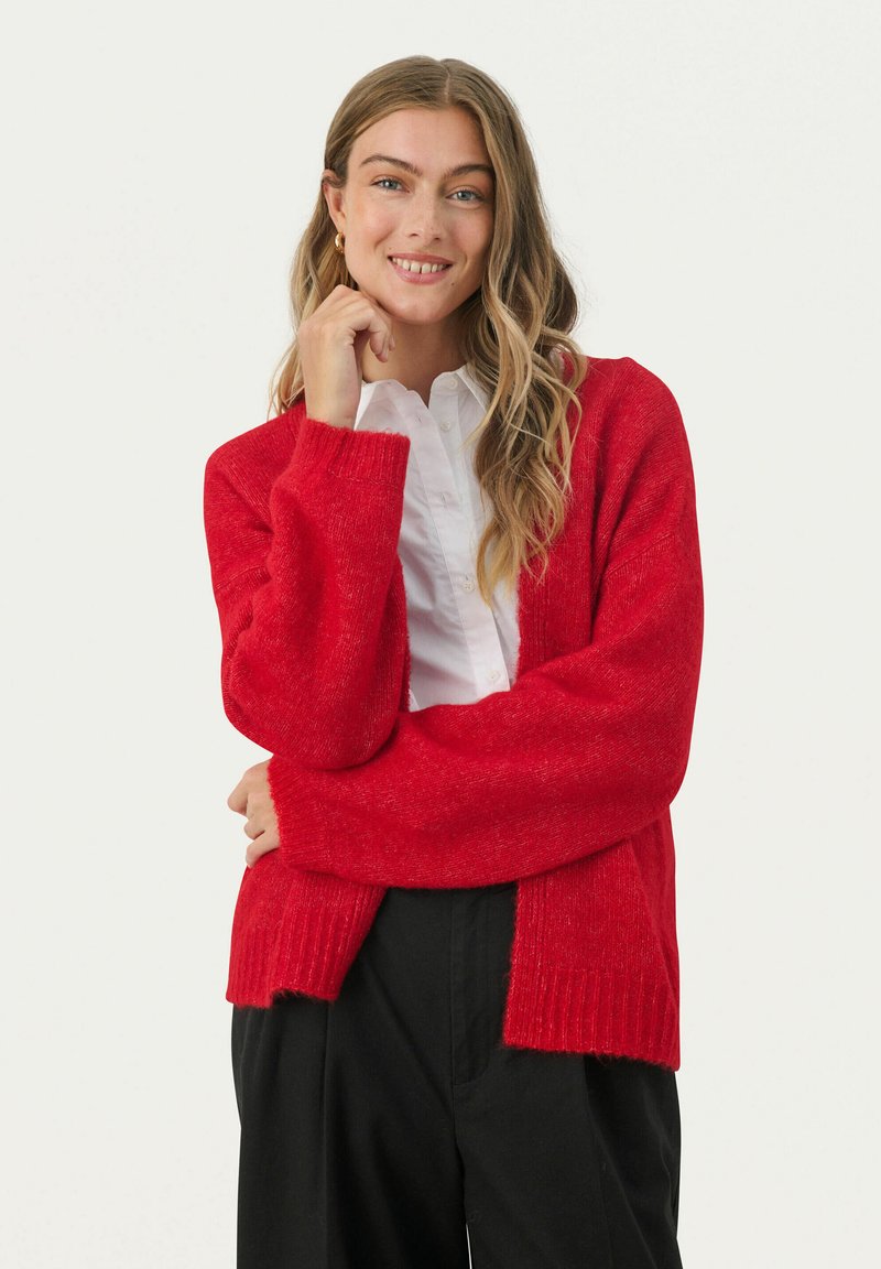 Červený pletený cardigan s mäkkou textúrou, otvoreným predným dielom a dlhými rukávmi. Žebrované manžety a spodný lem pridávajú detail. Spárované s bielou košeľou a čiernymi nohavicami.