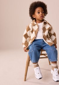 Braune und cremefarbene kariertes Fleecejacke, weißes Hemd, blaue locker sitzende Jeans, beige Socken, weiße Turnschuhe, sitzend auf einem Holzstuhl.