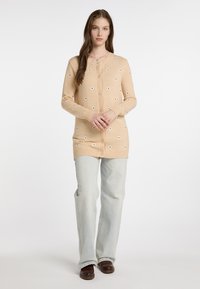 Beige gestrickte Strickjacke mit Knopfleiste, ausgestattet mit weißer floralem Stickerei, gerippten Bündchen und elongated Design, kombiniert mit hellblauen Jeans.