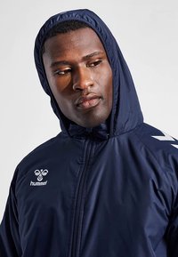 Chaqueta con capucha azul marino hecha de un material liso y ligero. Cuenta con una cremallera frontal, logo en el pecho y detalles blancos en los hombros.