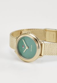 Gold mesh strap watch with a round green face featuring a subtle sparkle, gold hour and minute hands, and a small logo accent.