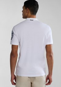 Bílá polo košile s krátkým rukávem, vyrobená z bavlny, má logovou nášivku na levém rameni a standardní design límce.