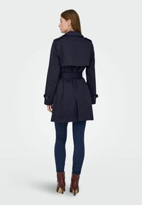Femme aux cheveux bruns raides portant un trench-coat bleu marine avec ceinture, un jean skinny bleu foncé et des bottines marron, vue de dos sur un fond uni.