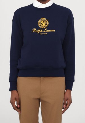 Person iført navy sweatshirt med guld Ralph Lauren New York våbenskjold logo, hvid skjorte med krave under og beige bukser.