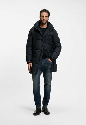 Mand iført en sort polstret vinterjakke, mørk sweater, blå jeans og sorte støvler står med den ene hånd i lommen.