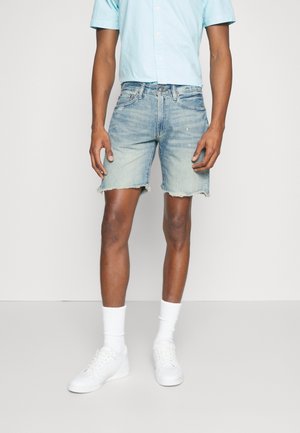 Persoon die lichtblauw shirt met korte mouwen draagt, lichte versleten spijkershorts met rafelige zoom, witte sportsokken en witte sneakers, staand.