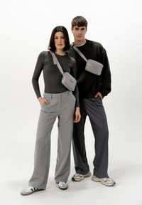 Šedé crossbody tašky nošené přes černé a šedé outfity; model má na sobě dlouhý rukáv a široké kalhoty s jemným vzorem.