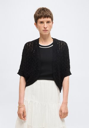 Kobieta o krótkich brązowych włosach nosi złote kolczyki koła, czarny krótki sweter na guziki z dzianiny nałożony na czarny top i białą warstwową spódnicę.