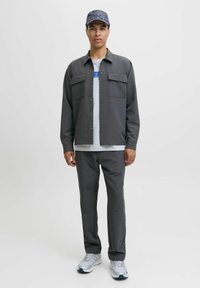 Jonge man staande in een donkergrijze jasje en broek, met witte sneakers, een wit shirt met een blauw patroon, en een gemêleerde donkergrijze pet.