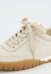 Beige suede sneaker featuring textured surface, slick round laces, gold accent at the lace tip, and a rubber sole with rounded grooves.