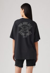 T-shirt noir oversize à manches courtes, avec un imprimé graphique blanc d'une planète et le texte "NOUS SOMMES TOUS DE LA POUSSIÈRE D'ÉTOILES" au dos.