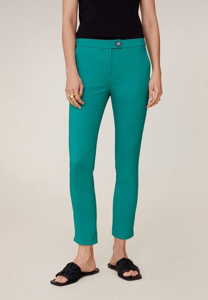 Femme portant un pantalon slim chevillé bleu sarcelle, un top noir sans manches, des sandales noires ouvertes à l'avant, et des bagues dorées aux deux mains.