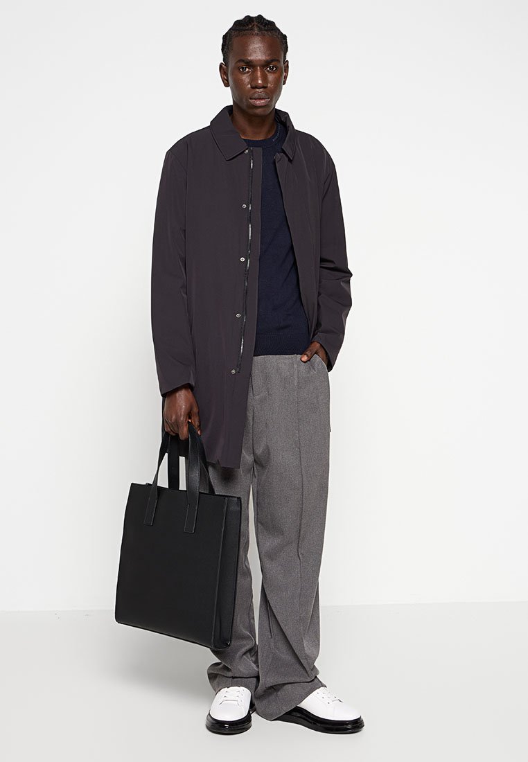 Czarny lekki płaszcz na granatowym swetrze, szare spodnie i białe buty. Trzyma czarną torbę na ramię. Proste, nieozdobne elementy designu.