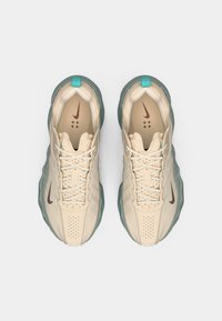 Beige atletiske sko med mesh-overdel og teal-accenter. Funktionen har et brunt logo, strukturerede såler og et ventileret tåområde. Snørebåndsdesign.