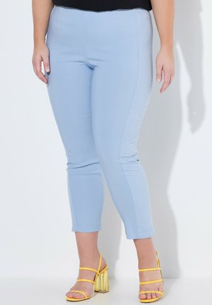 Kobieta w jasnoniebieskich dopasowanych spodniach do kostek i żółtych sandałach na wysokim obcasie stojąca przy białej ścianie.