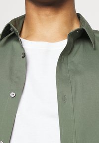 Close-up van een persoon die een witte ronde hals t-shirt draagt onder een opengemanierd olijfgroene overhemd.