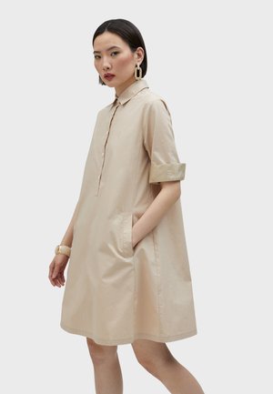 Vestido de camisa de algodón beige con cuello tipo solapa, mangas de tres cuartos, bolsillos laterales y un sutil detalle bordado. Textura suave.