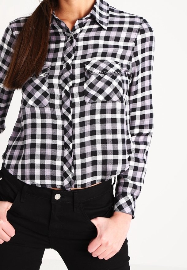 Donna che indossa una camicia a quadri bianca e nera con tasche anteriori e pantaloni neri, con le mani in tasca.