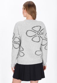 Hellgrauer Strickpullover mit schwarzen Schlaufenmustern. Mit Rundhalsausschnitt und gerippten Bündchen. Getragen zu einem schwarzen Faltenrock.