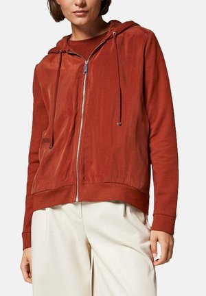 Kvinde iført rustrød lynlåshættetrøje med snore og off-white løse bukser, stående med afslappet holdning mod en hvid baggrund.