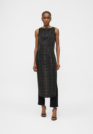 Nő fekete ujjatlan ruhában, amelyen függőleges, fehér pöttyös vonalak vannak, fekete nadrággal és fekete pántos magassarkúval, fehér háttér előtt állva.