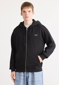 Čierna mikina z bavlnenej zmesi, so zapínaním na zips vpredu, klokanymi vreckami a malým bielym logom "HUGO" na ľavom hrudníku.