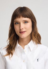 Camicia bianca con colletto classico, caratterizzata da un piccolo logo ricamato scuro sul lato sinistro del petto. Tessuto in cotone liscio.