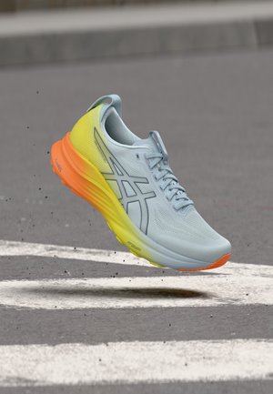 Chaussure de course bleu clair avec semelle jaune et orange, flottant au-dessus de l'asphalte avec des marquages routiers blancs.