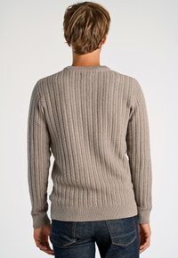 Beige stickad tröja med ribbad textur, rundad halsringning och tighta ärmar. Bakifrån visas en detaljerad kabelstickad design. Kombinerad med mörka jeans.