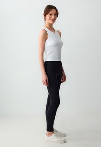 Top bianco senza maniche con texture a coste abbinato a leggings neri a vita alta; il modello indossa sneaker bianche, posando su uno sfondo neutro.